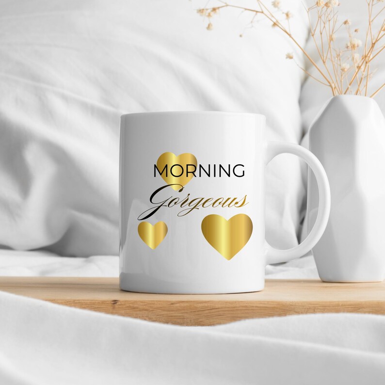 Op de afbeelding: Witte keramische mok met de woorden "MORNING Gorgeous" in zwart schrift en gouden hartaccenten. De mok heeft een gebogen handvat. Zachte, witte achtergrond.