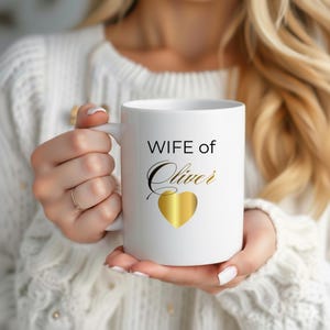 Op de afbeelding: Witte keramische mok met de tekst "WIFE of Oliver" in zwarte en gouden letters, met een gouden hart. De mok wordt vastgehouden door een persoon die een witte trui draagt.
