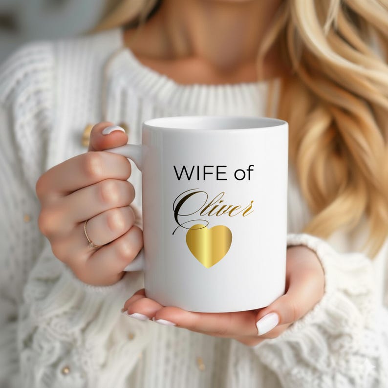 Op de afbeelding: Witte keramische mok met de tekst "WIFE of Oliver" in zwarte en gouden letters, met een gouden hart. De mok wordt vastgehouden door een persoon die een witte trui draagt.