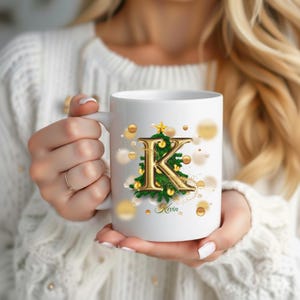 Peut inclure: Mug en céramique blanche avec la lettre "K" dorée ornée d'un sapin de Noël. L'arbre est orné de boules dorées et d'une étoile. Le nom "Kevin" est écrit en dessous. Des points dorés de bokeh entourent la tasse.