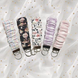 May include: Five fabric key fobs with metal hardware. The key fobs have different patterns: plaid, space-themed, white with pink stars, lavender, and pink. The key fobs are on a white background with gold stars.
