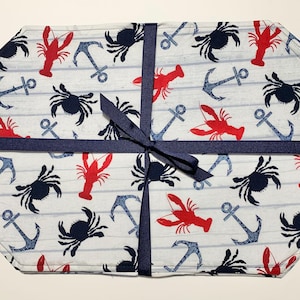 May include: A set of nautical-themed placemats featuring a pattern of red lobsters, navy blue crabs, and anchors on a white and light blue striped background. The placemats are tied with a navy blue ribbon.