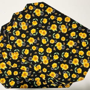 Könnte beinhalten: Vier achteckige Tischsets mit schwarzem Hintergrund. Die Tischsets haben ein sich wiederholendes Muster aus gelben Sonnenblumen, grauvioletten Blumen und kleinen Bienen. Die Ränder der Tischsets sind einfarbig schwarz.