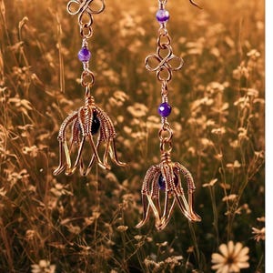 Puede incluir: Un par de pendientes hechos a mano de color dorado. Cada pendiente presenta un diseño floral con una cuenta morada y un dije decorativo de color dorado. Los pendientes cuelgan de un gancho dorado. El fondo es un campo borroso de flores silvestres.