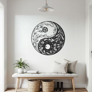 Puede incluir: Una obra de arte mural Yin y Yang en blanco y negro. El diseño presenta patrones florales y de hojas dentro de cada mitad del símbolo. La obra de arte está montada en una pared blanca sobre un banco con cojines, cestas y una planta en maceta.