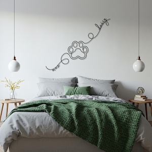 May include: A black metal wall art piece featuring a paw print design with the names "Max" and "Daisy" in cursive script. The artwork is mounted on a white wall above a bed with gray bedding and a green throw blanket.