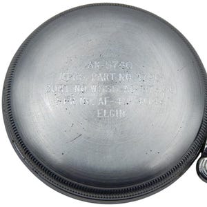 K&ouml;nnte beinhalten: Nahaufnahme eines silberfarbenen Uhrenr&uuml;ckens mit eingraviertem Text. Der Text lautet "AN-5740", "MFRS. PART NO. 1720", "CONT NO W535-AC-37880", "SER NO. AF-43-4045" und "ELGIN". Der Uhrenr&uuml;cken hat ein strukturiertes, kreisf&ouml;rmiges Design mit detailliertem Rand.