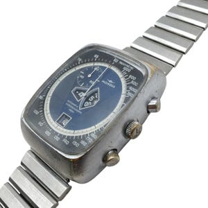 Op de afbeelding: Een vintage Mondia automatische chronograaf horloge met een vierkante, zilverkleurige kast en een blauwe wijzerplaat. Het horloge heeft een datumweergave, meerdere sub-wijzerplaten en een tachymeterschaal. Zilverkleurige, rekbare band.