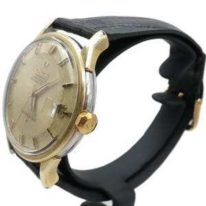 Könnte beinhalten: Eine gold- und silberfarbene Omega Constellation Uhr mit schwarzem Lederarmband. Das Zifferblatt der Uhr ist goldfarben mit goldenen Zeigern und Stundenmarkierungen. Die Uhr hat eine Datumsanzeige bei 3 Uhr. Die Uhr wird auf einem schwarzen Ständer präsentiert.