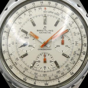 May include: A vintage Breitling Chronomat wristwatch with a silver case and a cream-colored dial. It has orange hands and sub-dials, with black numerals and markers. The watch face displays the Breitling logo and the word "Chronomat".