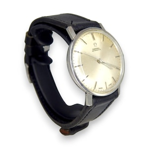 Puede incluir: Un reloj de pulsera Omega Automatic vintage con esfera plateada y agujas plateadas. El reloj tiene una correa de cuero negro. La esfera del reloj muestra el logotipo de Omega y la palabra "SWISS".