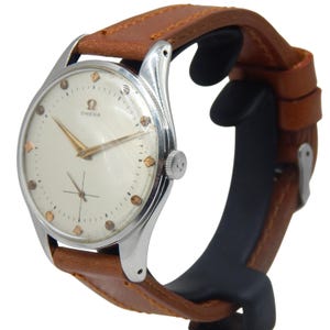 Può includere: Un orologio da polso Omega vintage con quadrante color crema, lancette dorate e indici delle ore. L'orologio ha una cassa in acciaio inossidabile e un cinturino in pelle marrone. L'orologio è esposto su un supporto nero.