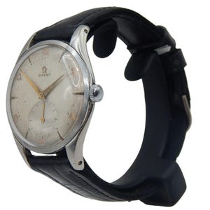 Puede incluir: Reloj de pulsera Omega vintage con caja plateada y correa de cuero negro. La esfera es blanquecina con agujas e indicadores dorados. El reloj tiene una subesfera y el logotipo de Omega.