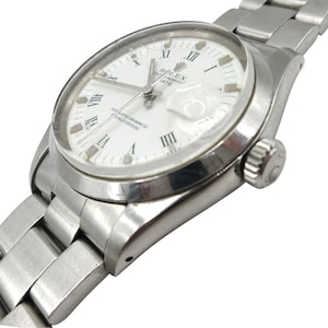 Puede incluir: Un reloj Rolex Oyster Perpetual Date de acero inoxidable con esfera blanca. El reloj presenta marcadores de hora con números romanos, una ventana de fecha a las 3 en punto y una pulsera plateada. Un reloj clásico.