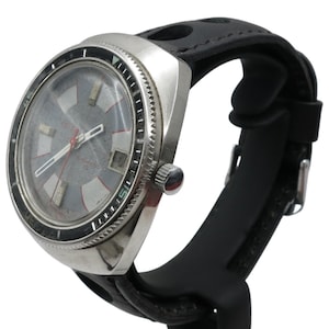 Puede incluir: Un reloj de pulsera vintage con caja plateada y correa de caucho negra. La esfera del reloj es gris con detalles en rojo y blanco. El reloj tiene un bisel giratorio y una corona lateral.