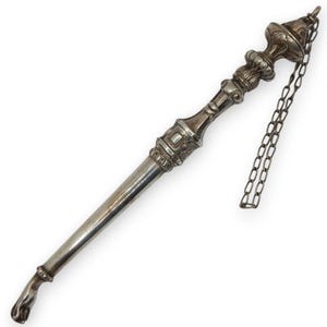 Puede incluir: Un objeto plateado y ornamentado con un mango y elementos decorativos. El objeto tiene un diseño detallado con una cadena adjunta en la parte superior. El mango tiene un extremo en forma de mano. El artículo parece ser de metal.