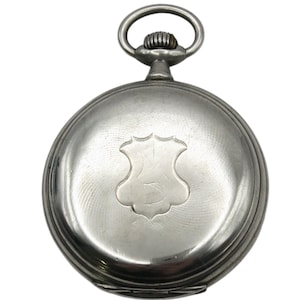 May include: A silver pocket watch with a circular face and a decorative shield emblem. The watch has a ring at the top for attaching a chain. The watch case is made of silver and has a ridged design around the edges.