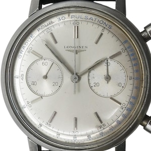 May include: Close-up of a vintage Longines chronograph watch. The watch has a silver face with two sub-dials, a seconds hand, and the text "GRADUE POUR 30 PULSATIONS". The watch has a stainless steel case and a black leather strap.