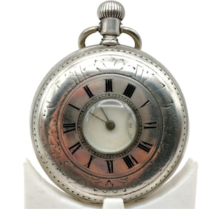 Puede incluir: Un reloj de bolsillo plateado con esfera blanca y números romanos negros. El reloj tiene un pequeño dial de segundos y grabados decorativos alrededor del bisel. La corona y el asa están en la parte superior. El reloj se exhibe sobre un soporte blanco.