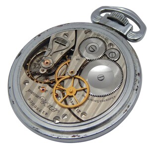 K&ouml;nnte beinhalten: Eine silberne Taschenuhr mit ge&ouml;ffneter R&uuml;ckseite, die die komplizierten Innenmechanismen zeigt. Das Zifferblatt zeigt Zahnr&auml;der, Federn und den Text "B.W. Raymond" und "21 Jewels". Die Uhr hat eine silberne Schlaufe zum Anbringen einer Kette.
