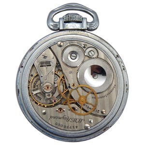 K&ouml;nnte beinhalten: Eine silberfarbene Taschenuhr mit offenem Zifferblatt und sichtbaren Zahnr&auml;dern und Mechanismen. Das Zifferblatt zeigt die Marke Elgin TL Watch Co. S.A., den Namen B.W. Raymond und die Nummer 41756006. Die Uhr hat eine silberne Aufzugswelle und eine Metallschlaufe.