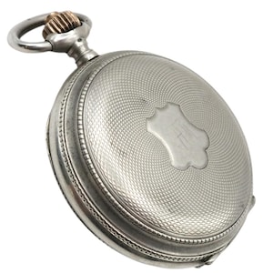 Puede incluir: Reloj de bolsillo plateado con esfera texturizada y un elemento decorativo en forma de escudo. El reloj tiene un lazo de cadena plateado y una corona de cuerda dorada. Un reloj clásico.