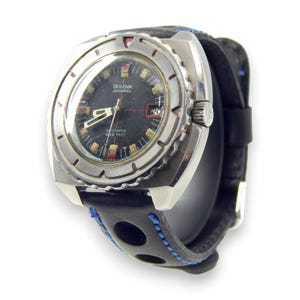 Puede incluir: Un reloj vintage Bulova Snorkel con esfera negra, bisel plateado y correa de cuero negro con costuras azules. La esfera del reloj muestra la fecha y las palabras "AUTOMATIC 666 FEET".