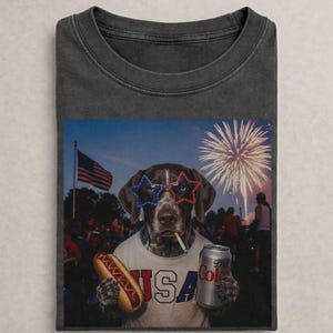 May include: A charcoal grey t-shirt featuring a graphic of a dog wearing star-shaped red, white, and blue glasses and a white t-shirt with the text USA. The dog holds a hot dog and a silver soda can against a backdrop of fireworks and a flag.