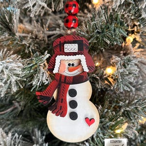 May include: Wooden snowman Christmas ornament with a red and black plaid hat and scarf. The snowman has a carrot nose, black button eyes, and a red heart. The hat says "BRRR". Two red and black beads hang above the snowman.