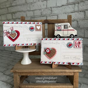 May include: Valentine's Day themed wooden postcards with red, white, and blue detailing. One postcard has a heart design and the text "Happy Valentine's Day." A small white Cupid Express van is also present.