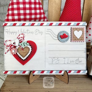 May include: A wooden Valentine's Day postcard with a red, white, and blue border. The postcard features a red heart with a wooden heart in the centre, the text "Happy Valentine's Day", and "P.S. I Love You". The postcard is propped up on a wooden stand.