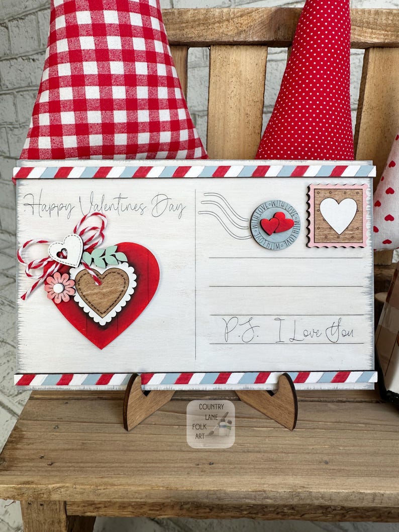 May include: A wooden Valentine's Day postcard with a red, white, and blue border. The postcard features a red heart with a wooden heart in the centre, the text "Happy Valentine's Day", and "P.S. I Love You". The postcard is propped up on a wooden stand.