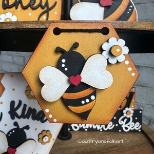 May include: A hexagonal wooden sign with a painted bee design. The bee is black and yellow with white wings and a red heart. The sign is painted yellow with white polka dots and has a white flower with a yellow center. The sign says "Summer Bee" at the bottom.