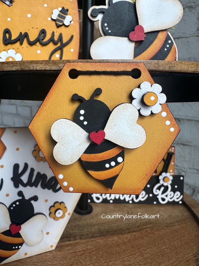 May include: A hexagonal wooden sign with a painted bee design. The bee is black and yellow with white wings and a red heart. The sign is painted yellow with white polka dots and has a white flower with a yellow center. The sign says "Summer Bee" at the bottom.