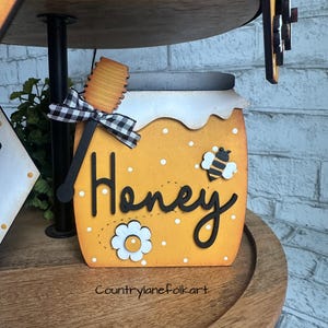 May include: A wooden honey jar with a black and white checkered bow, a wooden honey dipper, and the word "Honey" written in black. The jar is decorated with white polka dots and a small bee illustration. The jar is sitting on a wooden tiered tray.