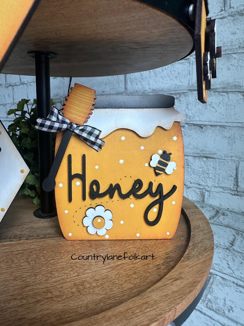 May include: A wooden honey jar with a black and white checkered bow, a wooden honey dipper, and the word "Honey" written in black. The jar is decorated with white polka dots and a small bee illustration. The jar is sitting on a wooden tiered tray.