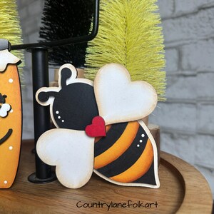 May include: A wooden bee ornament with black and yellow stripes, white heart-shaped wings, and a red heart on its body. The bee is sitting on a wooden surface.