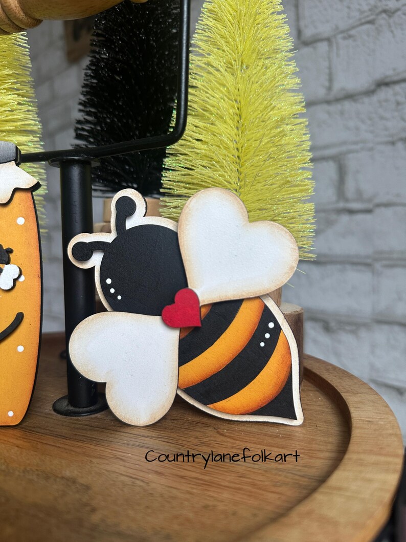 May include: A wooden bee ornament with black and yellow stripes, white heart-shaped wings, and a red heart on its body. The bee is sitting on a wooden surface.
