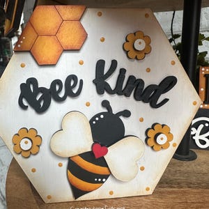 May include: A hexagonal wooden sign with a bee and honeycomb design. The sign says "Bee Kind" in black letters. The bee is yellow and black with white wings and a red heart.