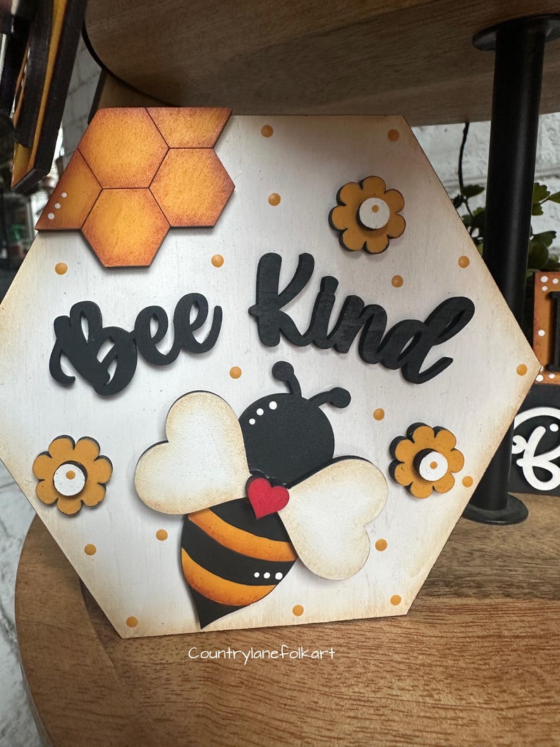 May include: A hexagonal wooden sign with a bee and honeycomb design. The sign says "Bee Kind" in black letters. The bee is yellow and black with white wings and a red heart.