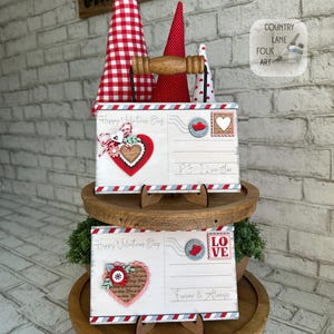 May include: Two Valentine's Day postcards with decorative hearts and the text "Happy Valentine's Day" and "Love". The postcards are displayed on a wooden tiered stand, with red and white cone-shaped decorations in the background.