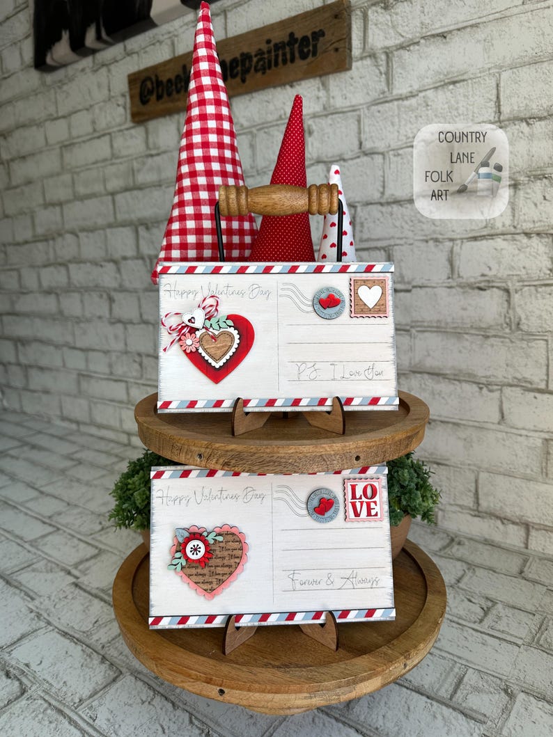 May include: Two Valentine's Day postcards with decorative hearts and the text "Happy Valentine's Day" and "Love". The postcards are displayed on a wooden tiered stand, with red and white cone-shaped decorations in the background.