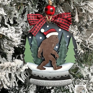 May include: A round Christmas ornament featuring a brown Bigfoot figure wearing a Santa hat, set within a snow globe design. The ornament has a black background with green trees and the text "MERRY SQUATCHMAS". It is adorned with a red and black plaid bow.