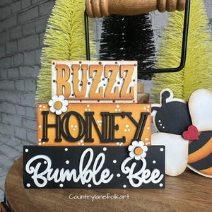 May include: A three-tiered wooden sign with the words "Buzz Honey Bumble Bee" in black and white lettering. The sign is decorated with white polka dots and white daisies. The sign is displayed on a wooden table with a black metal handle.