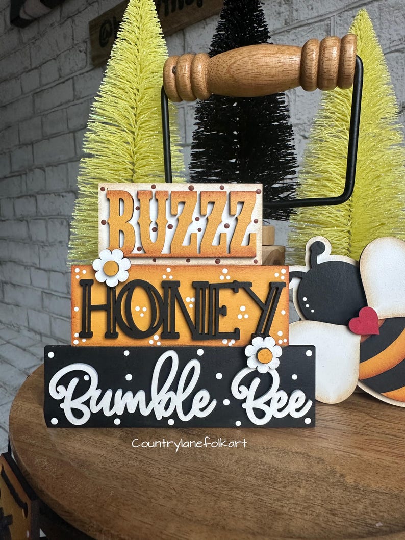 May include: A three-tiered wooden sign with the words "Buzz Honey Bumble Bee" in black and white lettering. The sign is decorated with white polka dots and white daisies. The sign is displayed on a wooden table with a black metal handle.