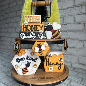 May include: A tiered tray decorated with bee-themed wooden signs. The signs feature phrases like "Buzz Honey", "Bumble Bee", "Bee Kind", and "Honey". The tray is also decorated with small wooden bees and honeycomb patterns.