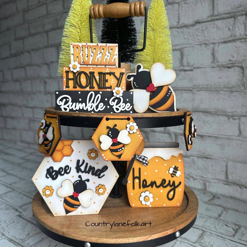 May include: A tiered tray decorated with bee-themed wooden signs. The signs feature phrases like "Buzz Honey", "Bumble Bee", "Bee Kind", and "Honey". The tray is also decorated with small wooden bees and honeycomb patterns.