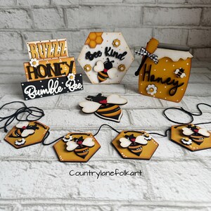 May include: Wooden bee-themed decorations with the words "Buzz Honey Bumble Bee", "Bee Kind", and "Honey". A garland of six hexagonal wooden bee ornaments with black and yellow bees and white flowers is included.