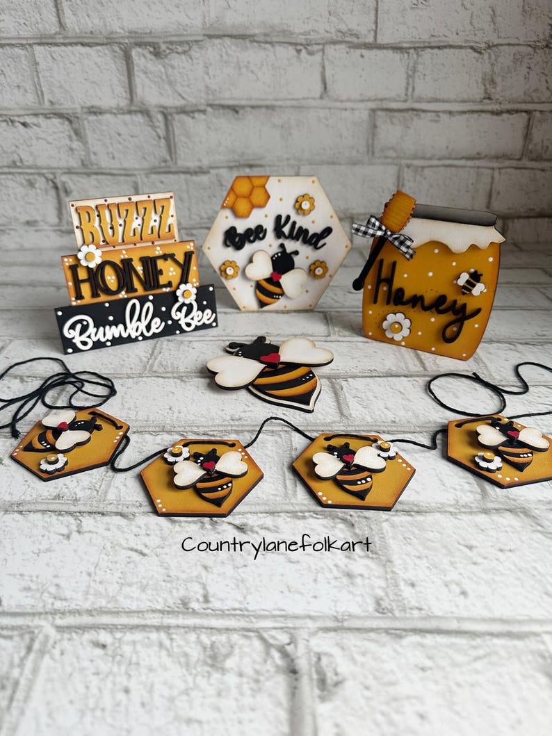 May include: Wooden bee-themed decorations with the words "Buzz Honey Bumble Bee", "Bee Kind", and "Honey". A garland of six hexagonal wooden bee ornaments with black and yellow bees and white flowers is included.