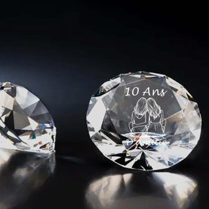 Peut inclure: Deux ornements en cristal clair en forme de diamant. L'un des ornements porte le texte "10 Ans D'amitié" et un graphique de deux personnes qui s'embrassent. L'autre ornement est flou. Le fond est sombre.